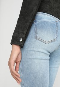 Sorte ruskindjakke med sølvtrykknapper ved håndleddene, parret med lyseblå denim jeans med en standard baglomme-design.