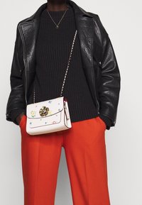 Weiße Leder-Crossbody-Tasche mit floralem Stickmuster, goldenen Akzenten und einer Kettenriemen. Getragen mit einem schwarzen Strickpullover und orangefarbenen Hosen.