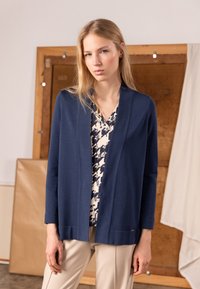 Mörkblå kofta med mjuk textur, öppen framkant och långa ärmar, lager-på-lager över en mönstrad blus, kombinerad med ljusbeige byxor.