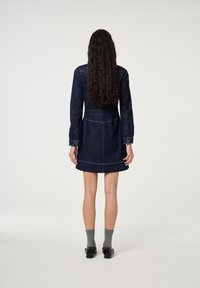Vrouw met lang krullend haar, gekleed in een donkere denim jurk, grijze sokken en zwarte schoenen, staand met haar rug naar de kijker tegen een effen achtergrond.