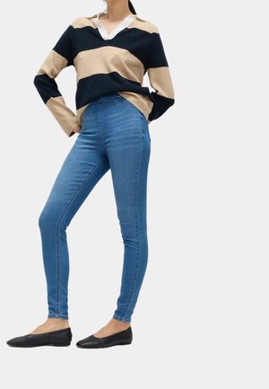 Kvinde iført blå skinny jeans, sort og beige stribet sweater med udskårede skuldre og sorte flade sko, stående med hænderne på hofterne.