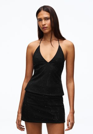 Femme aux longs cheveux foncés portant un top noir à col halter et une jupe courte assortie avec un motif texturé, debout sur un fond blanc uni.