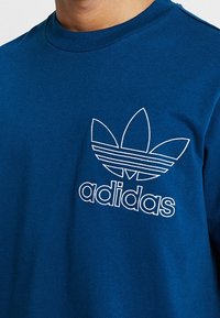 Blå bomulls T-shirt med en vit broderad Adidas-logotyp och trefoil-design på vänster bröst. Rund hals, korta ärmar, avslappnad passform.