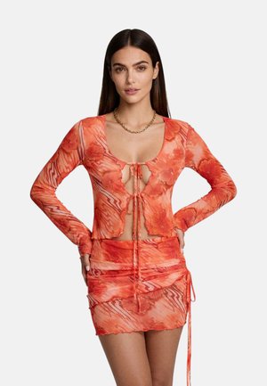 Donna che indossa un top arancione floreale trasparente a maniche lunghe con lacci sul davanti e una minigonna arricciata abbinata, su uno sfondo uniforme.