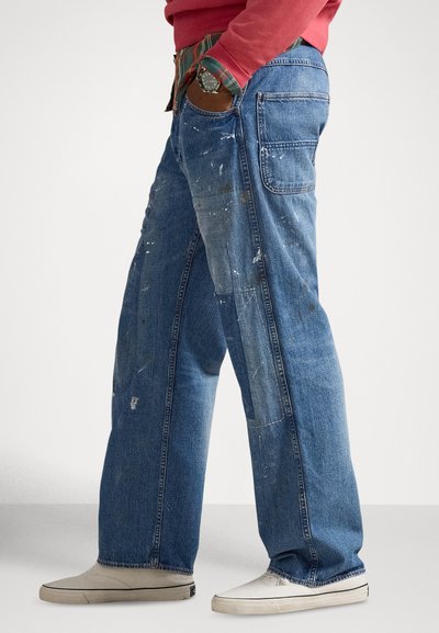 Blå denimjeans med en løs passform, med malingsprut, slitte områder og to baklommer. Kombinert med lyse joggesko.