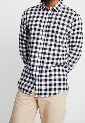 Homme portant une chemise à manches longues à carreaux noirs et blancs boutonnée, un pantalon beige, les mains derrière le dos, le torse visible sur un fond uni.