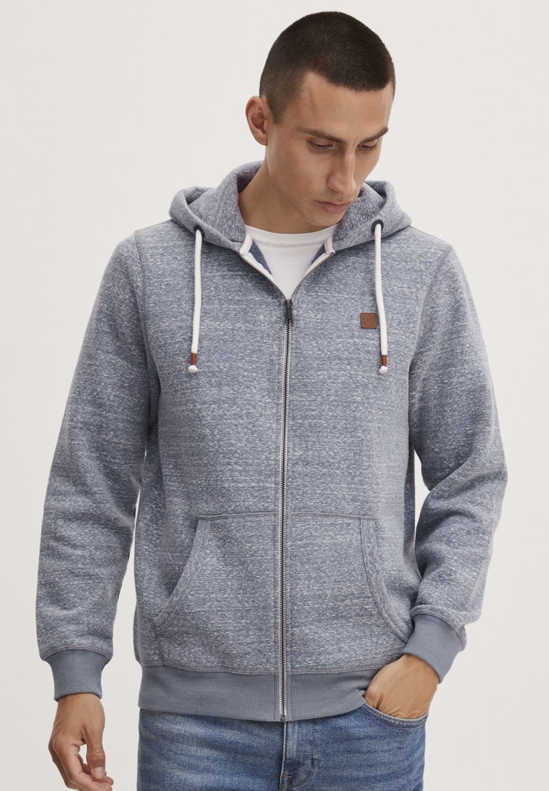 Felpa grigia con zip in tessuto strutturato, con cappuccio regolabile, tasche frontali e etichetta in pelle marrone sul petto. Si adatta a uno stile casual.