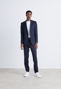 Suit navy con un design su misura, abbinato a una T-shirt bianca e scarpe bianche. Texture liscia e vestibilità moderna. Sfondo chiaro e styling semplice.