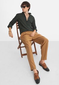 Camisa de botones a rayas verdes, pantalones beige y mocasines marrones con acentos negros. Modelo sentado en una silla de madera frente a un fondo neutro.
