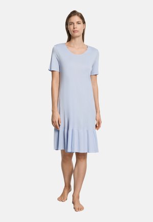 Nainen seisoo paljain jaloin, yllään vaaleansininen, lyhythihainen polvipituinen mekko, jossa on röyhelöreuna, yksiväristä valkoista taustaa vasten.