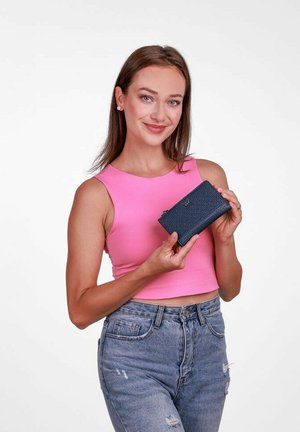 Námořnická modrá peněženka s texturovanou kůží, na zip a malým logem, kterou drží osoba oblečená v růžovém topu a světlých džínách.