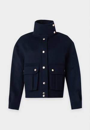 Femme portant une veste noire boutonnée avec de grandes poches, un jean gris clair avec revers, et des ballerines noires, debout devant un fond uni.
