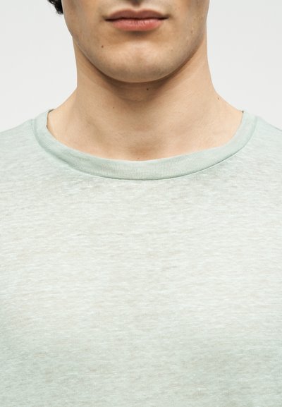 Εγγύς εικόνα του λαιμού και του κάτω μέρους του προσώπου ενός ατόμου που φοράει ανοιχτό γκρι μπλούζα με στρογγυλό λαιμό, μπροστά σε απλό φόντο.