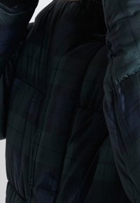 Person som bär en mörk rutmönstrad pufferjacka, justerar huvan, med fokus på jackans textur och färgmönster.