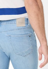 Jeans en denim bleu clair avec une texture lisse, des coutures visibles et une étiquette en cuir beige portant la mention "Brax" sur la ceinture.