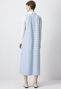 Femeie îmbrăcată cu o rochie midi fără mâneci, în carouri albastre deschise, peste o cămașă albă cu mânecă lungă, purtând pantofi de culoare crem, stând lângă un zid alb.