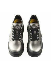 Zapatillas plateadas metálicas con cordones negros y una suela negra gruesa. Presenta un logotipo en la lengüeta y un forro interior amarillo.