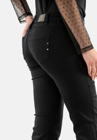 Pantalones vaqueros negros de denim con corte ajustado, con bolsillos traseros, herrajes plateados y una blusa larga de manga larga negra transparente texturizada con puntos de lunares.