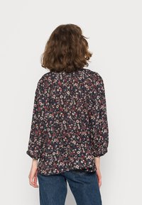 Femme aux cheveux bruns mi-longs portant un blouse à manches longues à fleurs sombres et un jean bleu, se tenant de dos devant un fond uni.