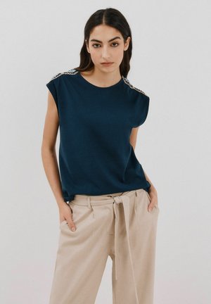 Haut bleu marine à manches courtes avec des embellissements distinctifs sur les épaules, associé à un pantalon taille haute beige clair avec un ruban à nouer à la taille.