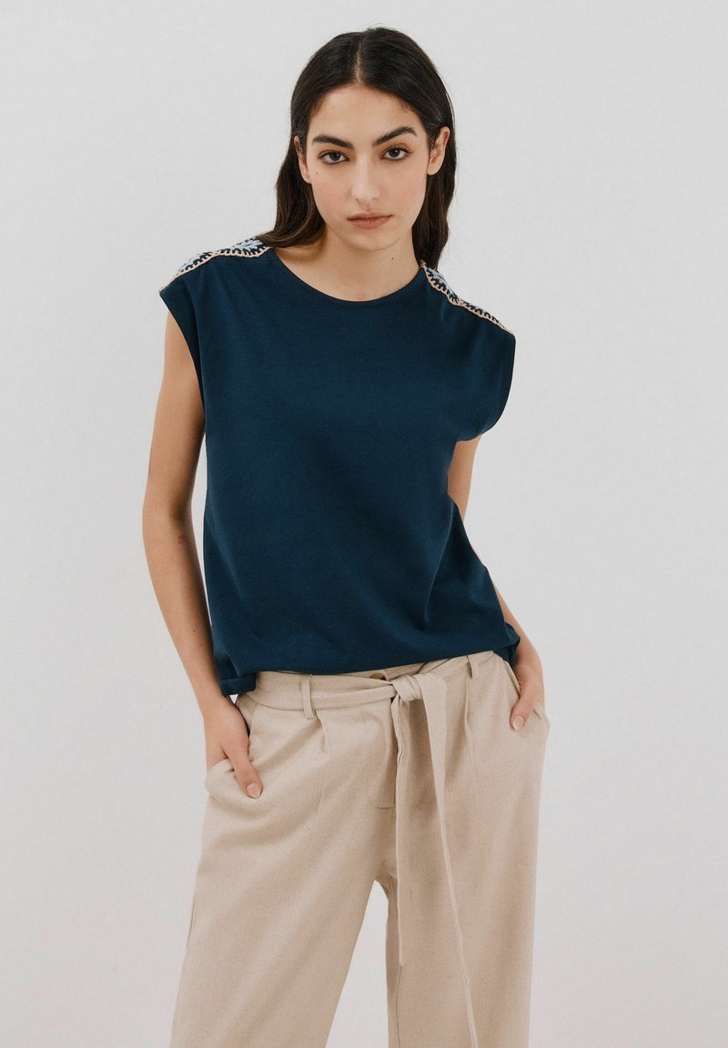 Dunkelblaues Kurzarmshirt mit auffälligen Schulterverzierungen, kombiniert mit hellbeigen, hoch taillierten Hosen mit gebundenem Bund.
