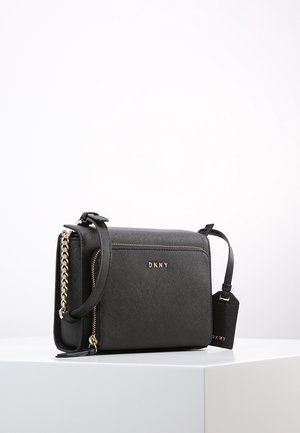 Sac bandoulière noir DKNY avec fermeture éclair dorée et détail chaîne, sangle réglable, et une étiquette noire assortie sur une surface blanche.