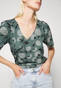 Top floral em verde com decote em V, mangas curtas bufantes e um padrão floral preto em tecido texturizado, combinado com calções de denim claros.