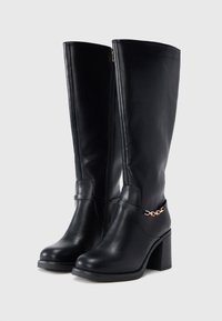 Μαύρες δερμάτινες μπότες μέχρι το γόνατο με χοντρό τακούνι, πλαϊνό φερμουάρ και λεπτό λουράκι με χρυσή αλυσίδα γύρω από τον αστράγαλο.