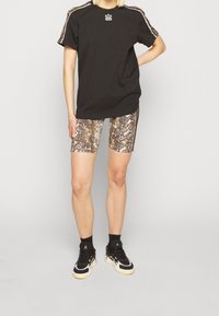 Henkilö, jolla on päällään musta Adidas-paita, jossa on painettuja raitoja ja logo, ruskeat käärmeennahkakuosiset pyöräilyshortsit, mustat sukat ja musta-valkoiset lenkkarit.