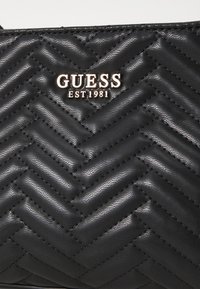 Sac à main noir matelassé avec un motif en chevron, arborant un logo métallique "GUESS EST 1981" en doré et une texture en cuir lisse.