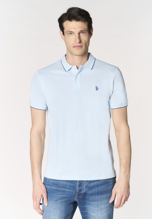 Giovane uomo che indossa una polo azzurro chiaro con rifiniture blu scuro e jeans blu, in piedi davanti a uno sfondo chiaro e semplice.