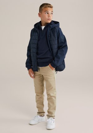 Marinblå puffjacka över en marinblå tröja, khakifärgade byxor och vita sneakers. Innehåller huva, dragkedjeddetaljer och texturerad tyg.