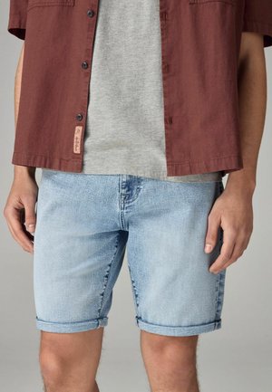 Lyseblå denimshorts med opstillede kanter, parret med en grå t-shirt under en burgunder kortærmet skjorte med sorte knapper og et mærke.
