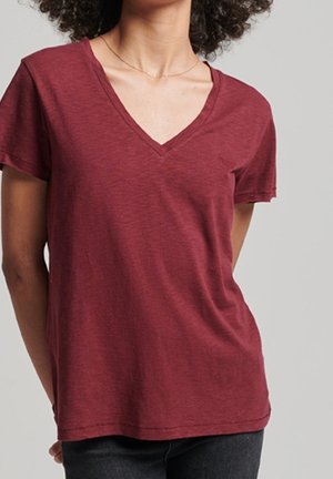 Kvinde iført en kortærmet, vinrød V-hals t-shirt med sorte bukser mod en ensfarvet grå baggrund.