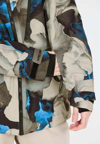 Hoodie in Beige, Blau und Schwarz mit abstraktem Muster. Mit Reißverschluss vorne, Klettverschluss an den Ärmeln und strukturiertem Stoff mit glatter Oberfläche.