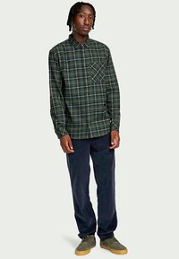 Camisa de botão xadrez verde, com bolso no peito, combinada com calças azul-marinho e sapatos cinza-marroados. Tecido suave com um ajuste casual.