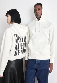 Due felpe bianche: una con una grande stampa nera "CALVIN KLEIN JEANS" sul retro, l'altra con un piccolo logo CK sul davanti. Entrambe sono in tessuto morbido.