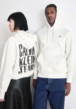 Due felpe bianche: una con una grande stampa nera "CALVIN KLEIN JEANS" sul retro, l'altra con un piccolo logo CK sul davanti. Entrambe sono in tessuto morbido.