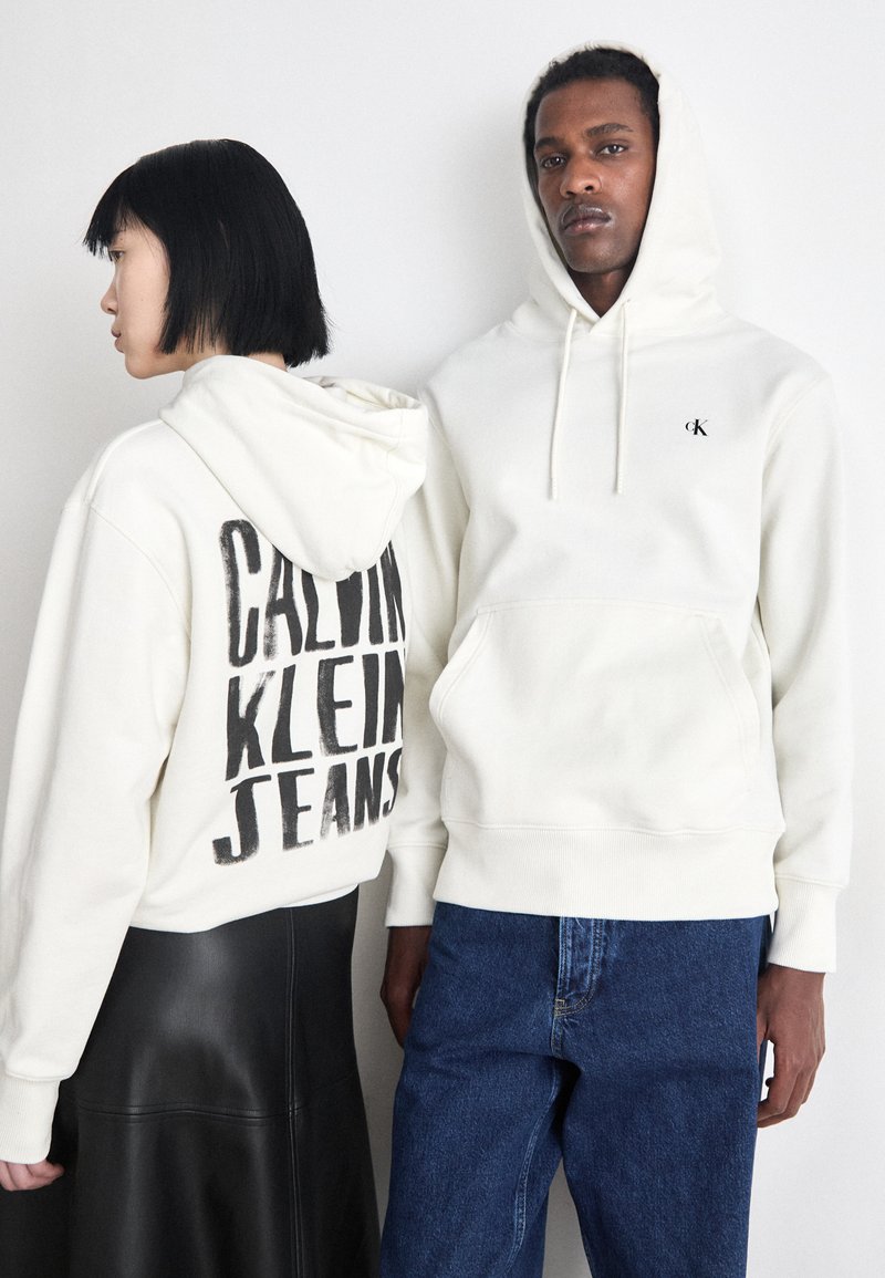 Due felpe bianche: una con una grande stampa nera "CALVIN KLEIN JEANS" sul retro, l'altra con un piccolo logo CK sul davanti. Entrambe sono in tessuto morbido.