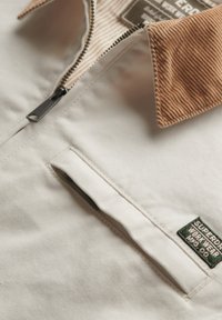 Hvid canvasjakke med kordfløjlskrage, lynlåslukning og en brystlomme med en grøn label. Tekstureret stof tilføjer visuel interesse.