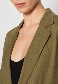 Olivgrön linnekavaj med slagkrage och avslappnad passform, lager-på-lager över en svart topp, som visar en texturerad tyg och rena linjer.