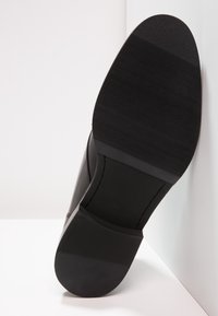 Černá podrážka pánské obuvi se strukturou na přední polovině, hladkým podpatkem a viditelným švem na bílém rohu.