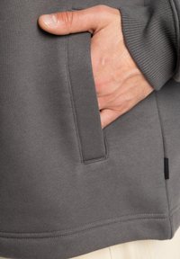 Tejido de sudadera gris con textura acanalada. Presenta un bolsillo lateral y una etiqueta negra cerca del dobladillo. Costuras visibles a lo largo de los bordes.