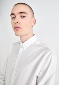 Chemise blanche à boutons en tissu lisse, dotée d'un col classique, d'une broderie de logo discrète et d'une coupe structurée.