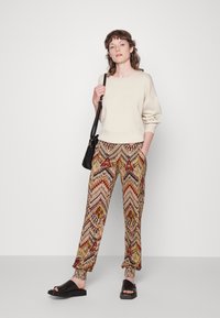 Sweat-shirt beige avec des épaules tombantes, associé à un pantalon à motifs dans des couleurs chaudes présentant des designs en zigzag et géométriques. Sandales à glisser noires.