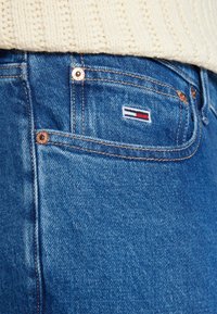 Modré džíny z denimu s malým vyšitým logem s červenými, bílými a modrými pruhy u zadní kapsy. Viditelné detaily švů.