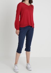 Blusa rossa con maniche lunghe e scollatura a V, abbinata a pantaloni blu navy a 3/4. Sneakers bianche con lacci completano l'outfit.
