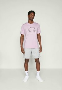 Tricou scurt cu mânecă de culoare violet deschis, cu logo și text negre, asortat cu pantaloni scurți gri cu model. Adidași și șosete albe completează ținuta.