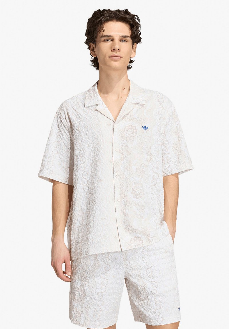 Giovane uomo che indossa una camicia bianca a maniche corte con bottoni e pantaloncini coordinati con un delicato motivo floreale strutturato e un logo blu sul petto della camicia.