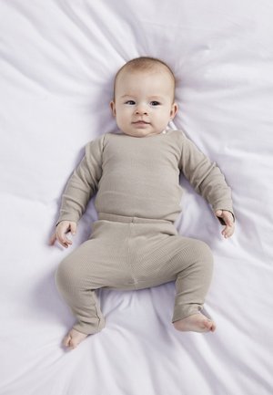 Completo beige a coste per neonati composto da due pezzi, con maniche lunghe, pantaloni aderenti e scollatura rotonda, posato su una leggera coperta lavanda.